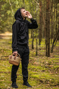 Man in black hooded clothing calling out while holding a woven basket in a sunlit woodland clearing with trees and mossy ground, outdoor forest portrait