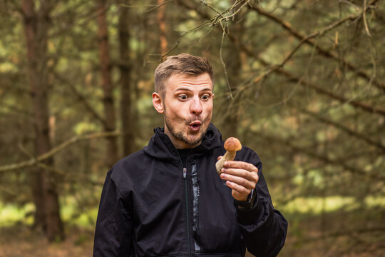 Surprised man holding a small brown mushroom while exploring a sunlit woodland, expressive face and dark outdoor jacket, forest nature portrait