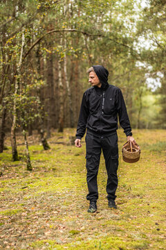 Man in black outdoor clothing carrying a woven basket while walking through a sunlit woodland with mossy ground and autumn foliage, vertical nature portrait
