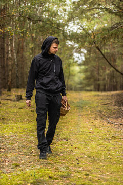 Hooded man carrying a woven basket while walking along a mossy forest path surrounded by trees and soft autumn foliage in a woodland landscape