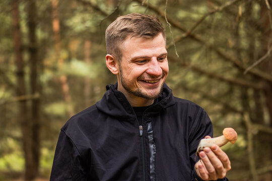 Smiling man holding a small porcini mushroom while standing in a wooded forest, wearing a black jacket during an outdoor nature walk, portrait photograph