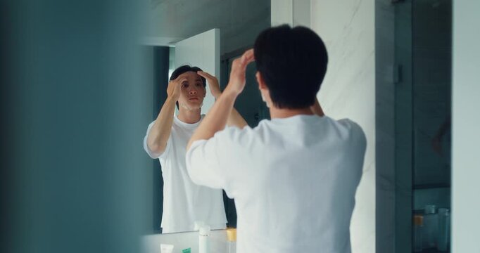 Young Asian man applies sheet mask to face in bathroom mirror at home. Male grooming skincare routine, Facial beauty treatment hydration, Daily health wellness moisturizer care concept.