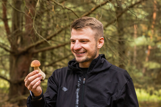 Smiling man holding a small mushroom while exploring a green woodland with mossy ground and dense trees, outdoor nature portrait in a forest