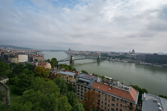 View of the Szechenyi Chain Bridge spanning the Danube River with historic city architecture and rooftops under a cloudy sky in Budapest, Hungary.