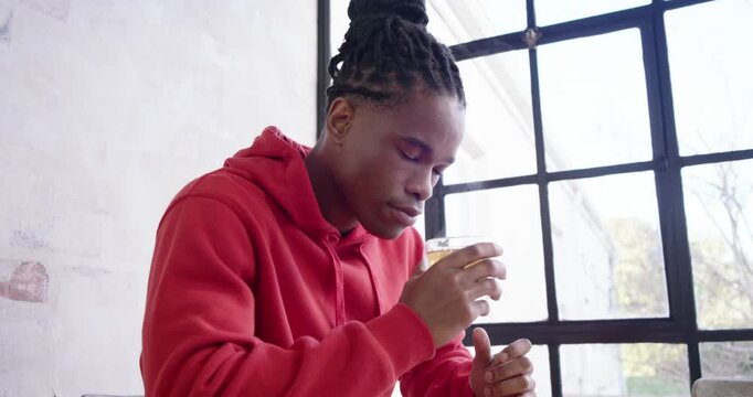 Man in red hoodie sitting at wooden table by grid window, sniffing steaming mug, sipping, thinking