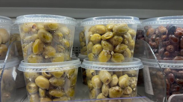 Packaged olives fill rows of chilled grocery display shelves. Transparent tubs hold green, brown and black olives in marinade, presenting varied deli assortment for meals, snacks and entertaining.