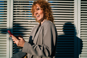 Profile portrait of joyful Caucasian female student holding smartphone against outdoor wall, celebrating instant mobile banking transfer confirmation cashback rewards and fund savings © BullRun