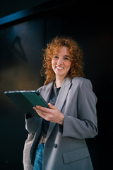 Portrait of smiling Caucasian female student looking at camera while holding digital tablet against black wall, celebrating e-commerce mobile retail application promotional discount sales © BullRun