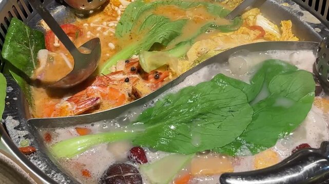 Close up footage of a divided hot pot simmering with shrimp, leafy greens, mushrooms and vegetables in two bubbling broths while a ladle moves through the soup.