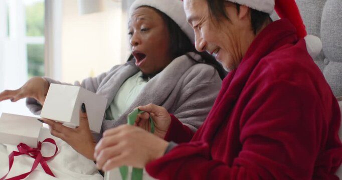 Senior man and woman in Santa hats on bed opening christmas gifts, pulling green ribbon laughing