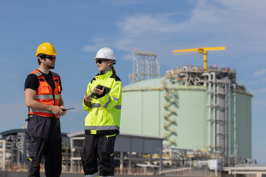 Workers in safety gear check data on tablet near LNG storage facility during day