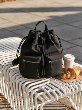 Backpack black leather zipper pocket street outdoor bench coffee croissant modern black backpack with zipper pocket detail white metal bench outdoor street setting beside takeaway coffee cup