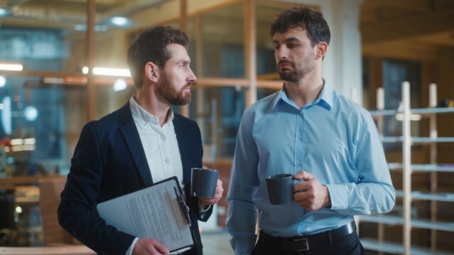 Two friendly calm Caucasian business men talking drinking coffee break pause in office. Relaxed businessmen male partners talk drink tea together communicating workers agents discussing corporate deal