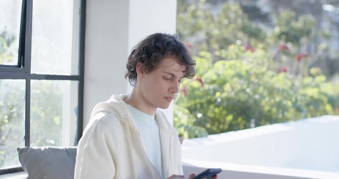 Adult male scrolling smartphone on cushioned bench by window, pausing and looking at garden plants