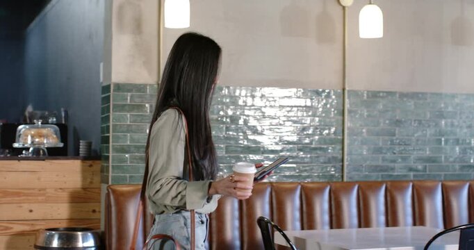 Asian woman entering from left, carrying coffee and device, scanning cafe and heading toward window