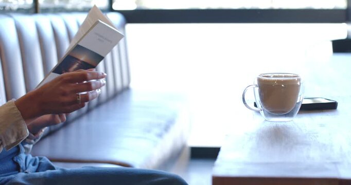 African American woman reading booklet, grasping glass mug, sipping at cafe booth