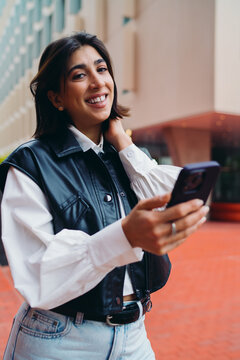 Hispanic woman user checking travel transport schedule smartphone app look at camera smile. Beautiful spanish female student typing mobile message near texturized modern building backdrop.