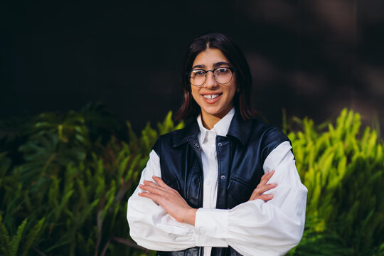 Confident spanish female standing with crossed arms smiling proudly against dark copy space wall backdrop. Happy hispanic woman student experiencing positive emotions in urban green park.
