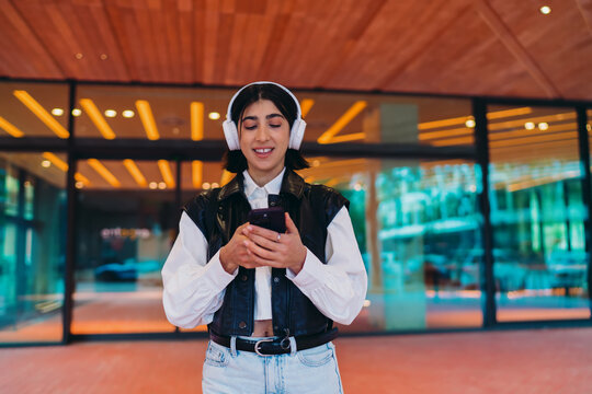 Hispanic woman streaming music playlist on mobile app via wireless headphones. Smiling spanish female using cellular network chat application over high speed 5G internet in urban location.