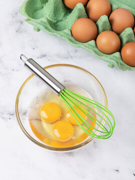 Whisk in glass bowl with cracked raw eggs and carton of brown eggs on marble table, top view