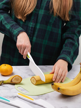 Teenage girl cutting banana with safe plastic knife on cutting board, close-up of hands without face, fruits on table
