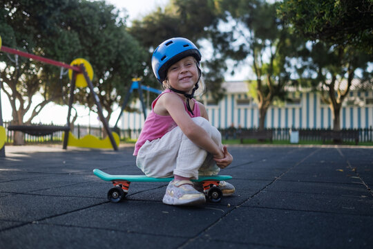 Cute Little Girl in Blue Helmet Sitting on Penny Board at Playground Outdoors
