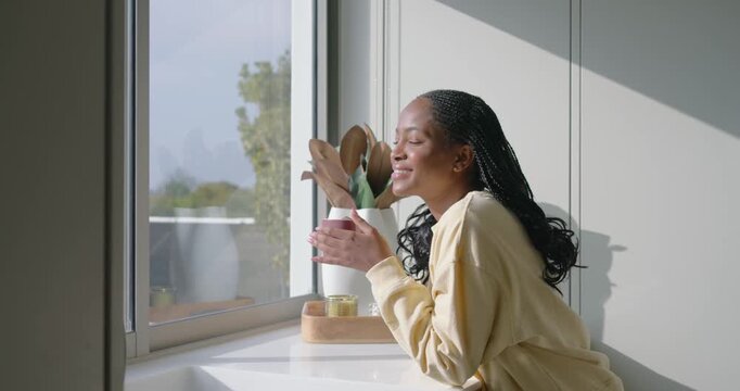 Adult African woman leaning kitchen windowsill holding mug, savoring, smiling, laughing as sunlight
