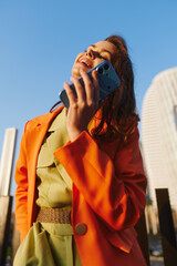 Fototapeta premium Happy young woman in an orange blazer laughs while raising her smartphone to her ear against a blue sky in the city. Joyful phone call moment. Concept for telecom and messaging app promotions.