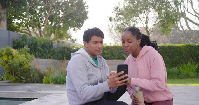 Couple wearing hoodies on yoga mat by pool, man raising black smartphone framing selfies for review