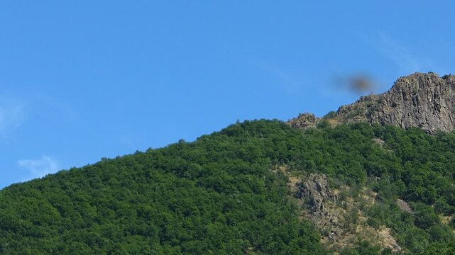 Rack focus from a hoverfly hovering in a blue sky to a rocky green mountain hill covered with forest
