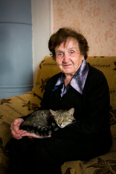 Portrait of an elderly woman sitting on a sofa and holding a sleeping tabby cat in her lap at home