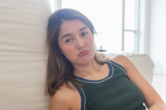 Young Asian woman pouting on a sofa, looking sad or bored indoors.