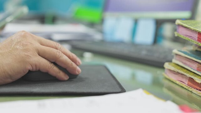 Employee hand moves computer mouse resting upon black mouse pad inside office workstation, managing daily workflow beside keyboard, stacked ledger notebooks, paper sticky notes, monitor display.