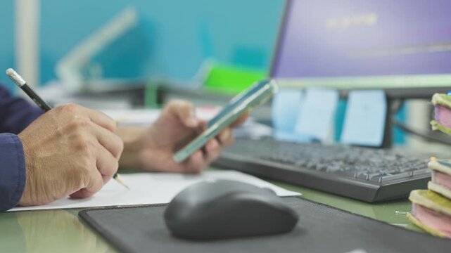 Focused office employee writes paper notes, holds mobile phone beside keyboard, computer monitor, desk accessories, managing corporate workflow, financial planning, administrative tasks.