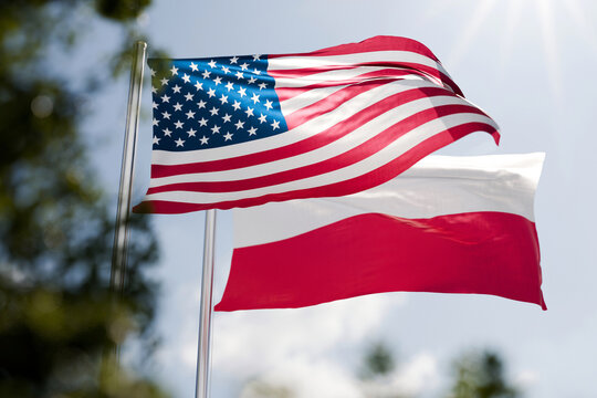 American and Polish national flags waving on flagpoles against a sunny blue sky. Concept of international diplomacy, partnership, bilateral relations, alliance, cooperation between USA and Poland.