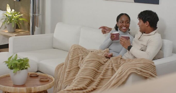 African American couple on sofa she pointing after joke he holding her sharing warmth sipping mugs