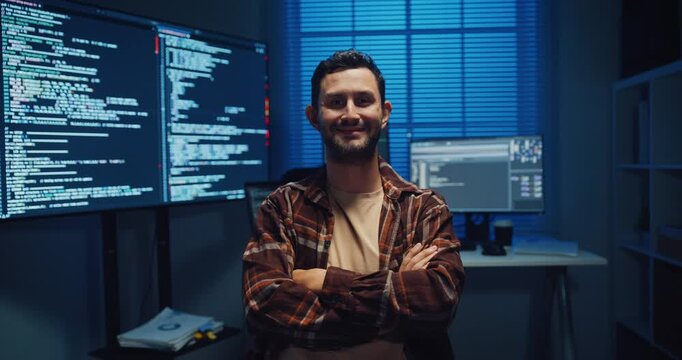 Portrait of confident Caucasian male programmer smiles looking at camera in office at night. Professional developer workspace, Secure computer network system, Advanced cybersecurity data concept.