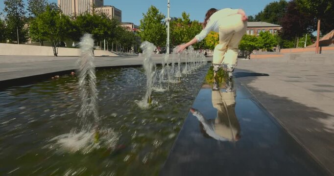 Young woman rollerblades beside city fountains as low tracking shot captures speed and adventurous energy. Urban sport, active lifestyle and summer recreation concept.