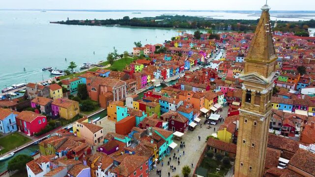 Aerial view of the colorful houses and the leaning bell tower of Chiesa Parrocchiale di San Martino di Castello in Burano with people walking along the canals in Venezia, Veneto, Italy.