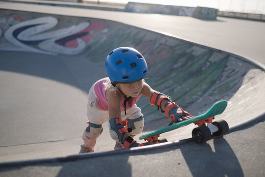 Determined Little Girl in Blue Helmet and Protective Pads Climbing Ramp with Skateboard at Skatepark