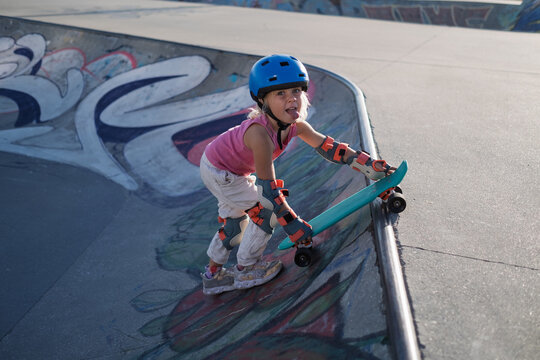 Determined Little Girl in Blue Helmet and Protective Pads Climbing Ramp with Skateboard at Skatepark