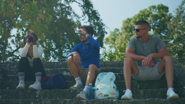 Three friends take a break on stone steps during an outdoor fitness activity. They are enjoying conversation and refreshments in a natural setting.