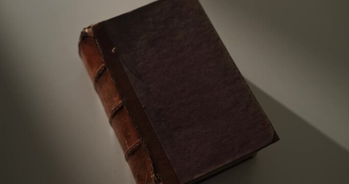 Cinematic orbit around an old vintage book with a worn leather spine resting on a white surface, illuminated by atmospheric local light and casting long dramatic shadows. Education, history, knowledge