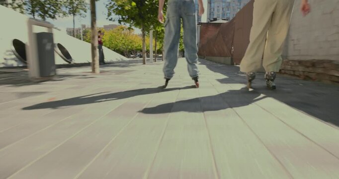 Two female friends rollerblade together along urban walkway as close tracking shot captures playful speed. Youth friendship, street sport and active lifestyle concept.