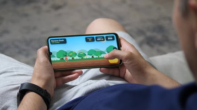 Young man plays a colorful side-scrolling runner on a smartphone, jumping over obstacles and collecting fruit in a fast, casual mobile gaming challenge.
