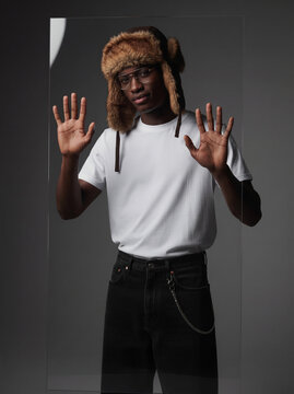Young Black man in white blank t-shirt, fur trapper hat and glasses posing with hands up behind glass, studio fashion portrait