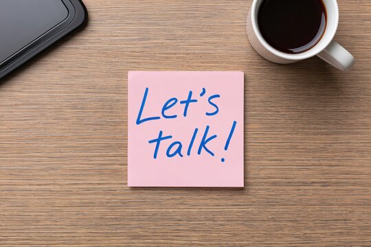 Note with a lets talk message on a pink sticky paper placed on a wooden office desk with coffee