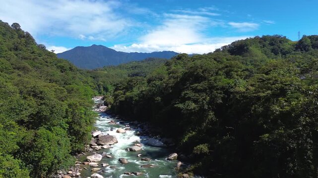 Fast-flowing turquoise river winds through a rocky tropical forest valley with distant mountains beneath a bright blue sky. The scenic landscape conveys adventure, freshwater ecology and nature travel