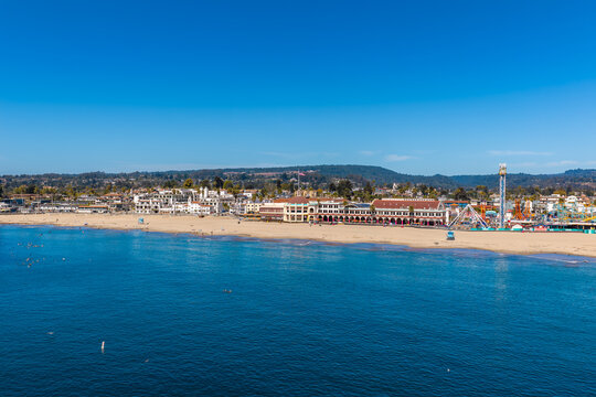 Aerial shot of Santa Cruz Beach Boardwalk along the sandy Pacific coast with ocean water, amusement park rides, and coastal buildings under a clear sky.