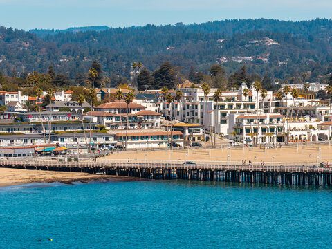 Aerial shot of Santa Cruz coastline in California featuring the wharf, sandy beach, coastal hotels, palm trees, and forested mountains under clear daylight.